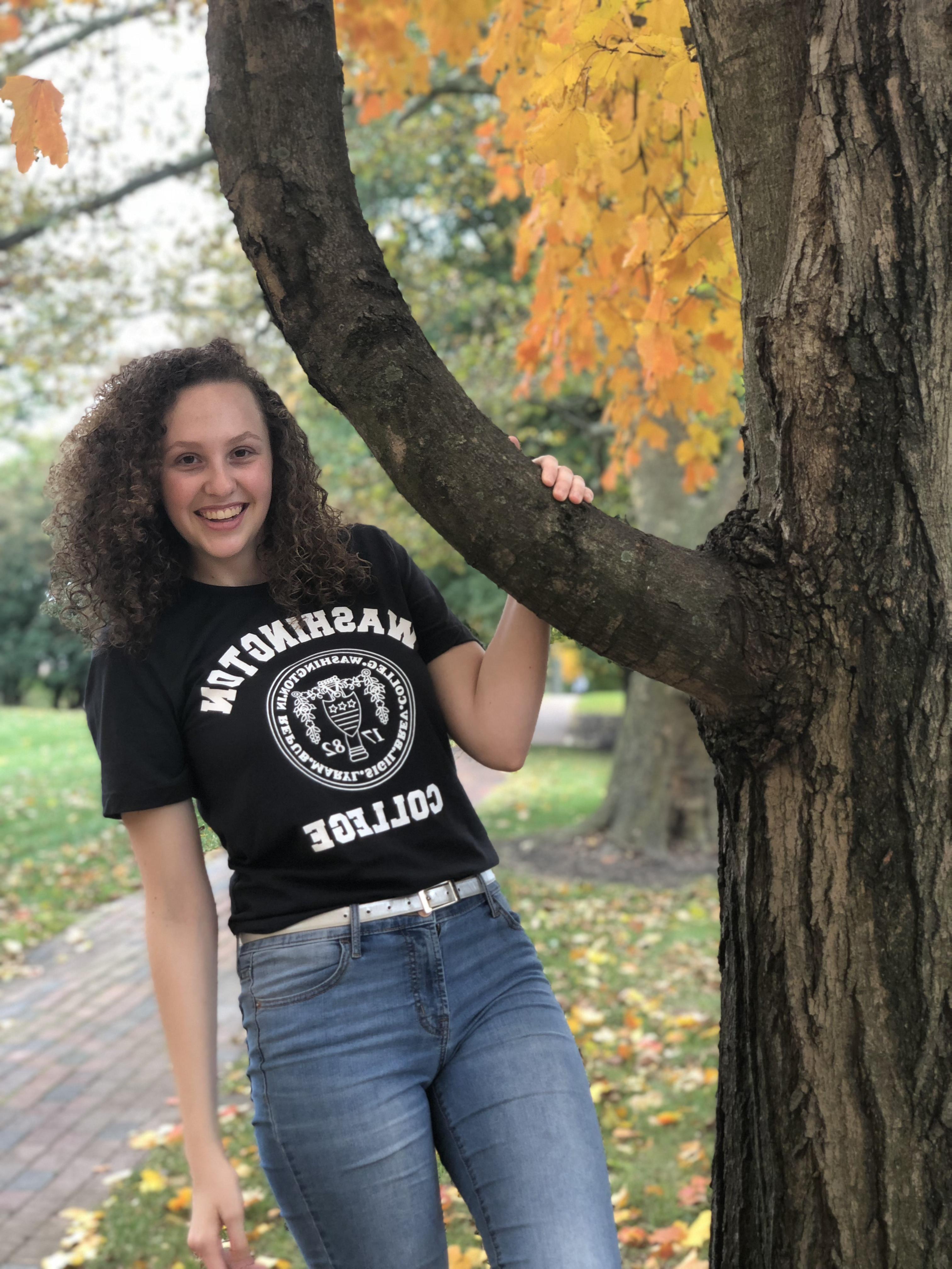 Carlee, smiling and wearing a black t-shirt with the Washington College logo, blue jeans, and a white belt in front of fall foliage. 